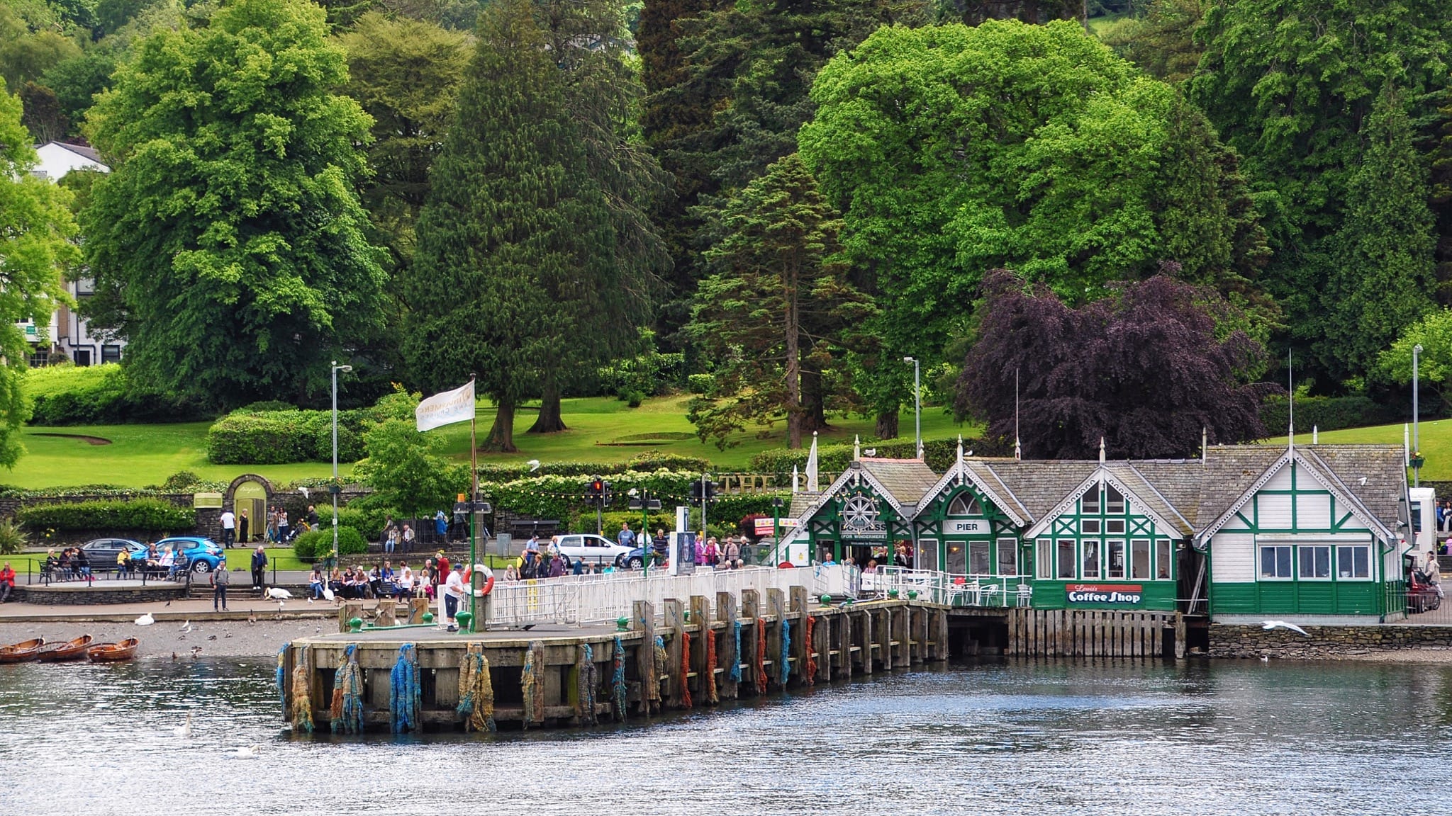 A view of Bowness Pier from the lake of Windermere, with green leafy trees behind it. Photo by Nilfanion, licensed CC-BY-SA-4.0.